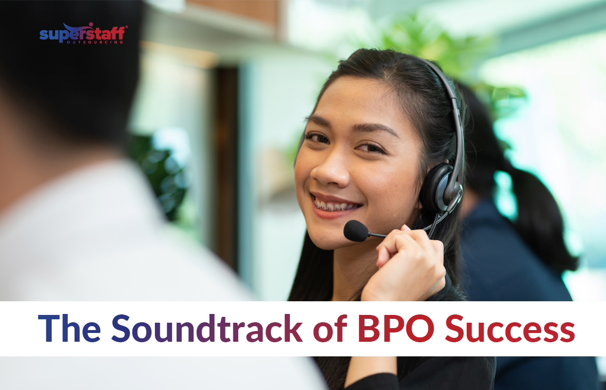 The-Soundtrack-of-BPO-Success_Featured A friendly female call center agent with long dark hair and braces, wearing a headset and smiling while assisting a customer. She is seated in a modern, well-lit call center environment with other agents working in the background. The SuperStaff Outsourcing logo is in the upper left corner. The text overlay reads, 'The Soundtrack of BPO Success,' with 'Soundtrack' in bold blue and 'BPO Success' in bold red, emphasizing the role of call center professionals in delivering excellent customer service.